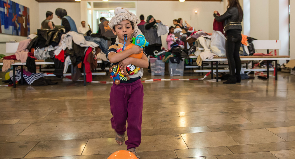 Ein kleiner Junge aus Syrien spielt in Stuttgart in der Kapelle Martinus-Haus mit einem Lufballon während er die Arme voll gespendeter Spielsachen hält (Bild: © dpa). 