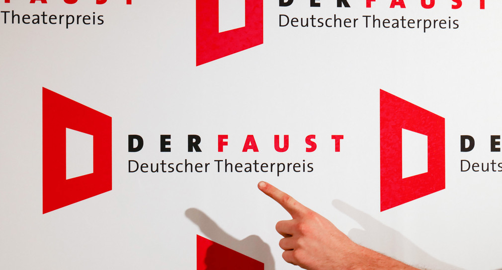 Eine Hand zeigt mit ausgestrecktem Zeigefinger auf das Logo mit dem Schriftzug „Deutscher Theaterpreis – DER FAUST“. (Foto: © dpa)