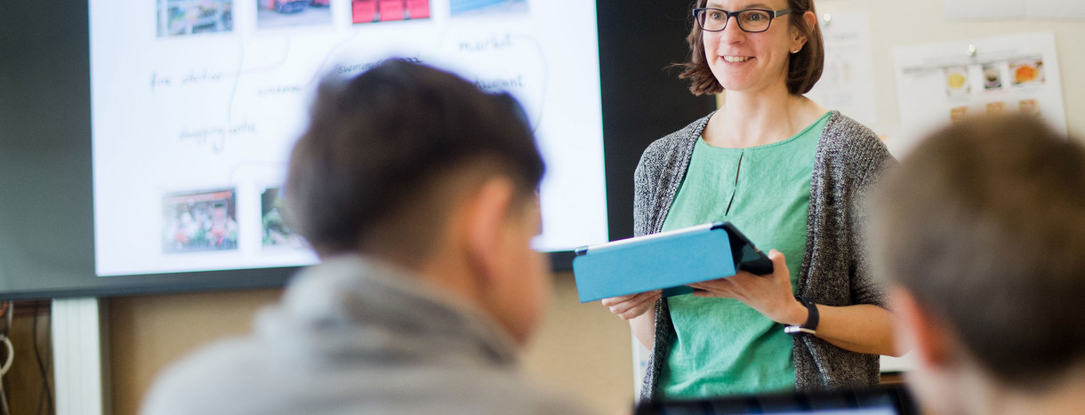 Eine Lehrerin mit einem Tablet in der Hand steht vor einer digitalen Tafel.