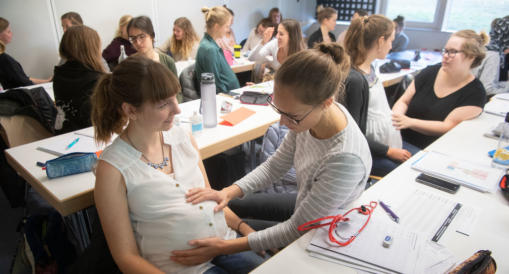 Eine Studentin der Hebammenwissenschaft hat während einer Übung in einer Vorlesung im Ausbildungszentrum DocLab des Universitätsklinikums Tübingen ein Kleidungsstück unter ihr Oberteil gestopft, um eine schwangere Frau darzustellen, damit eine Kommilitonin eine Betreuungsssituation mit ihr üben kann. (Bild: © dpa)