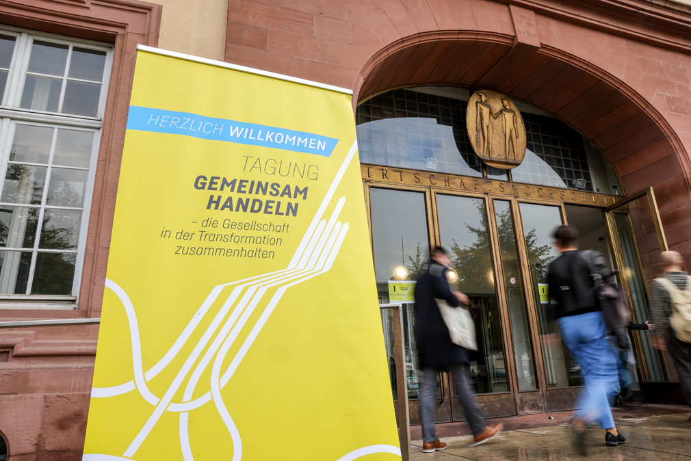 Eingang zur Tagung in der Universität Mannheim mit einem Banner mit der Aufschrift „HERZLICH WILLKOMMEN - TAGUNG GEMEINSAM HANDELN – die Gesellschaft in der Transformation zusammenhalten“.  