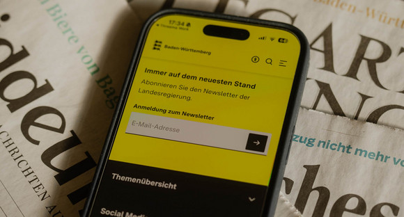 Ein Mobiltelefon zeigt die Anmeldung zum Newsletter auf dem Landesportal Baden-Württemberg.de. Darunter liegen drei Tageszeitungen.