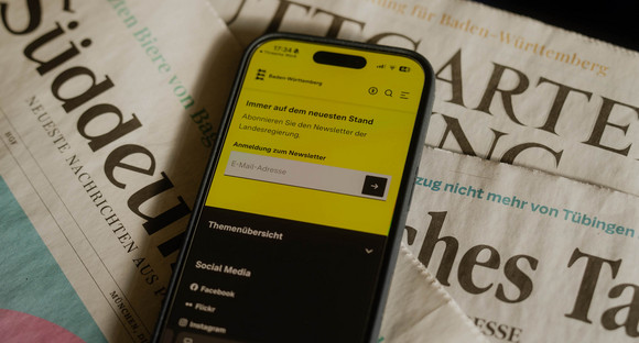 Ein Mobiltelefon zeigt die Anmeldung zum Newsletter auf dem Landesportal Baden-Württemberg.de. Darunter liegen drei Tageszeitungen.