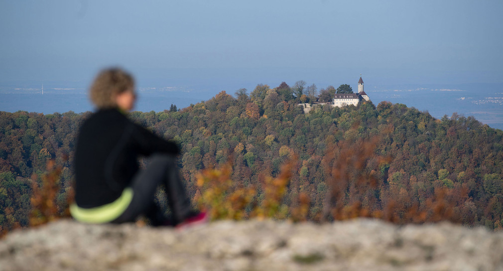 Eine Wanderin sitzt auf dem Breitenstein am Albtrauf der Schwäbischen Alb und betrachtet die Aussicht, im Hintergrund ist der mit herbstlichem Wald bedeckte Teckberg zu sehen, auf dessen Gipfel die Burg Teck thront. 