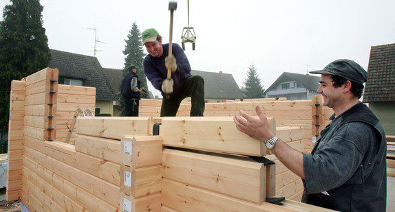 Zwei Bauarbeiter fügen beim Bau eines Holzhauses vorgefertigte Balken zu einer Hauswand aufeinander. (Foto: © dpa)