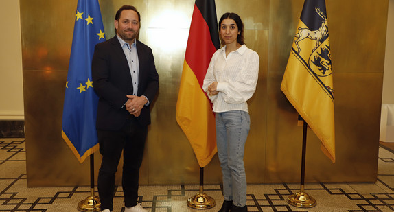 Staatssekretär Florian Hassler (links) und Friedensnobelpreisträgerin Nadia Murad (rechts) stehen in der Villa Reitzenstein in Stuttgart vor Fahnen.