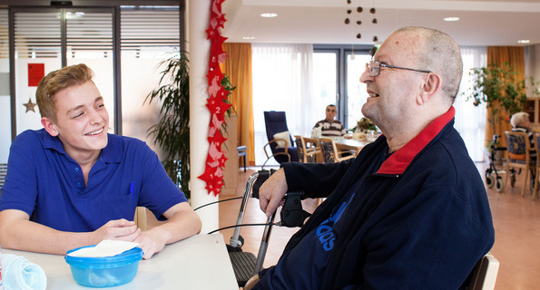 An einem Tisch im Pflegeheim ist ein junger Pfleger im Gespräch mit einem älteren Mann.