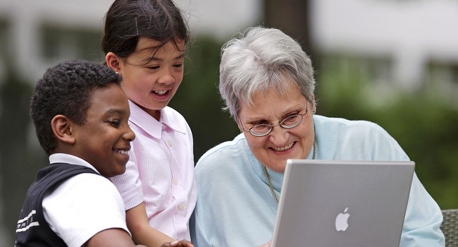Eine Seniorin arbeitet in einer Seniorentagesstätte zusammen mit Kindern an einem Laptop (Bild: © dpa).