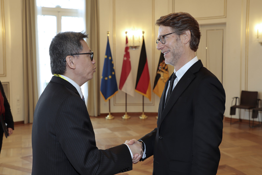 Staatsminister Dr. Florian Stegmann (rechts) begrüßt den Botschafter der Republik Singapur, Lee Chong Hock (links). Im Hintergrund stehen Fahnen.