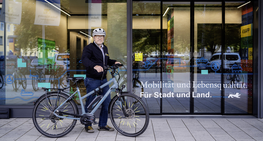 Verkehrsminister Winfried Hermann trägt einen Fahrradhelm und steht mit Fahrrad vor dem Eingang zum Verkehrsministerium.