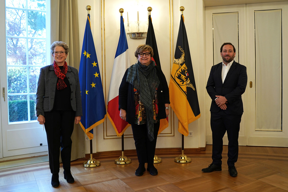 von links nach rechts: Staatsrätin Barbara Bosch, Brigitte Torloting, Vizepräsidentin der Region Grand Est, und Staatssekretär Florian Hassler stehen vor Fahnen.