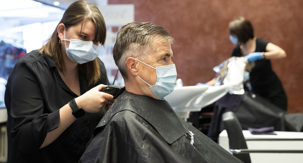Eine Friseurin eines Salons schneidet einem Kunden die Haare. Beide tragen einen Mundschutz. 