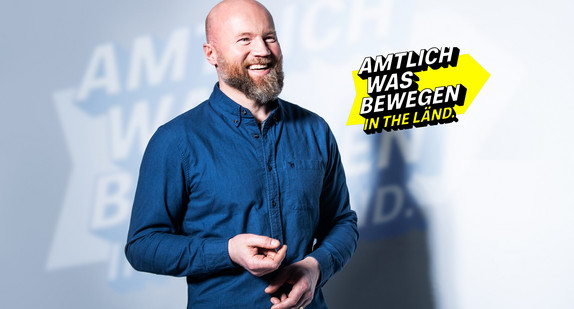 Lächelnder Mann in blauem Hemd vor dem Slogan "Amtlich was bewegen in THE LÄND."