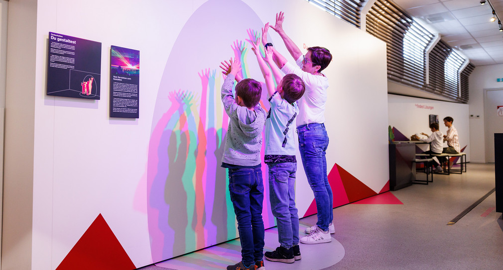 Eine Frau und zwei Jungen erzeugen an einem Exponat der experimenta mit ihren Armen farbige Schatten. 