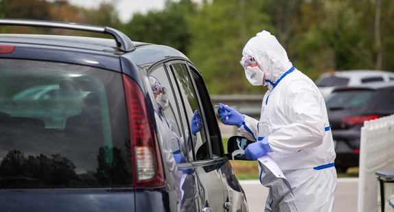 Eine Person im Schutzanzug steht am geöffneten Fenster eines Autos.