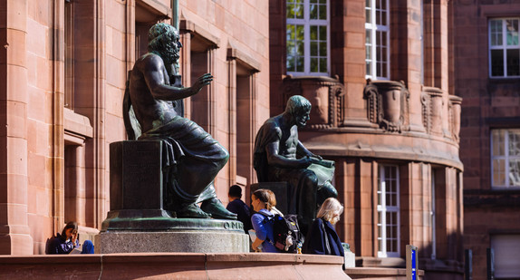Skulpturen von Homer (links) und Aristoteles stehen vor dem Haupteingang des Kollegiengebäude I (KG1) der Universität Freiburg während Menschen dazwischen hindurchgehen.
