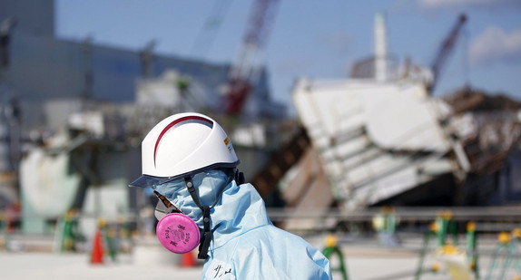 Ein Mann in Schutzkleidung und mit Atemmaske vor den Trümmern des zerstörten Atomkraftwerks in Fukushima, Japan (Bild: © Reuters, via dpa).