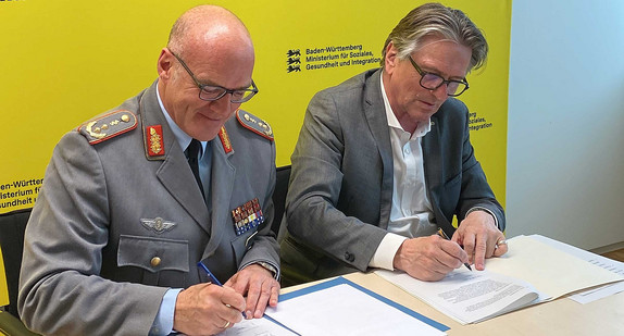 Der Kommandeur der Sanitätsakademie der Bundeswehr, Generalstabsarzt Dr. Hans-Ulrich Holtherm, und Gesundheitsminister Manne Lucha sitzen nebeneinander und unterzeichnen jeweils ein Dokument.