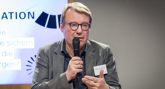 Man sieht Prof. Dr. Herbert Brücker in Großaufnahme. Hinter ihm sieht man einen Bildschirmausschnitt mit dem Key Visual der Veranstaltung. Herr Brücker hält ein Mikro in der Hand und wendet sich mit seiner Körperhaltung dem Publikum zu. Er trägt ein graues Jacket und ein dunkelblaues Hemd. Er ist Brillenträger