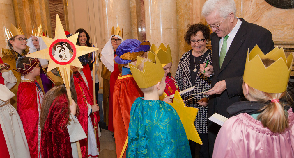 Ministerpräsident Winfried Kretschmann (r.) unterschreibt neben seiner Frau Gerlinde im neuen Schloss in Stuttgart Autogrammkarten für zahlreiche Sternsinger (Quelle: dpa).