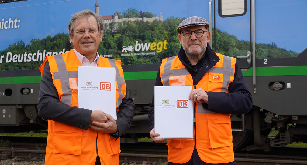 David Weltzien und Winfried Hermann stehen vor einem Zug und halten Mappen in die Kamera mit dem DB-Logo und dem Verkehrsministeriums-Logo bedruckt. Beide tragen eine orangene Warnweste.