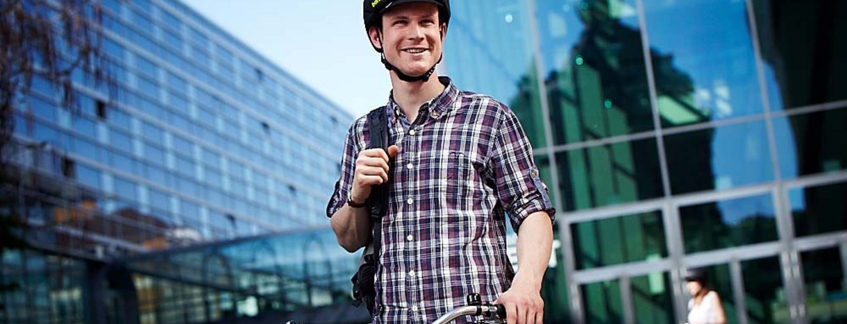Radfahrer in legerer Arbeitskleidung steht mit Rad und Helm vor einem Bürogebäude
