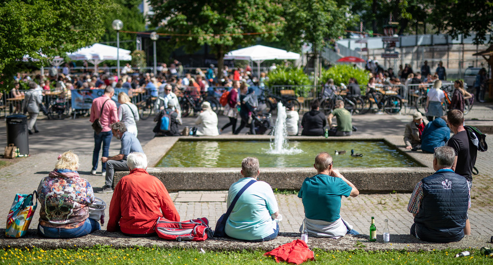 Menschen sitzen im Stuttgarter Schlosspark auf Mauer, im Hintergrund vollbesetzte Tische eines Biergartens.