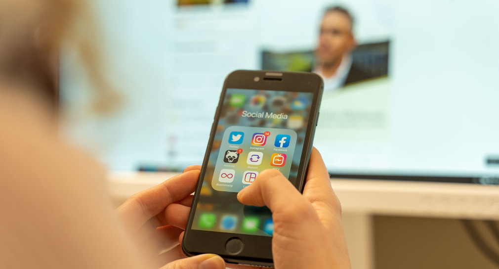 Jemand sitzt vor einem Computerbildschirm und hält ein Smartphone in der Hand auf dem mehrere Social-Media-Apps zu sehen sind.