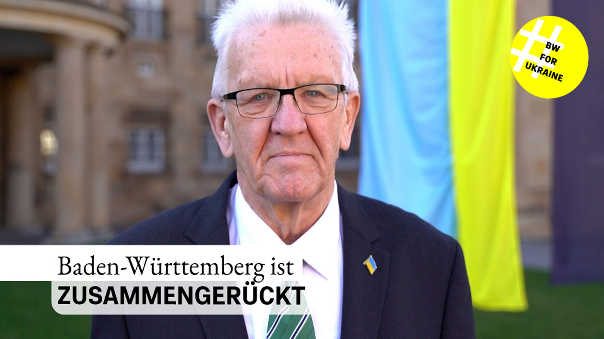 Ministerpräsident Winfried Kretschmann steht bei einer Ansprache zum Jahrestag des russischen Angriffskrieges gegen die Ukraine vor der Villa Reitzenstein in Stuttgart. Im Hintergrund ist die ukrainische Flagge zu sehen.