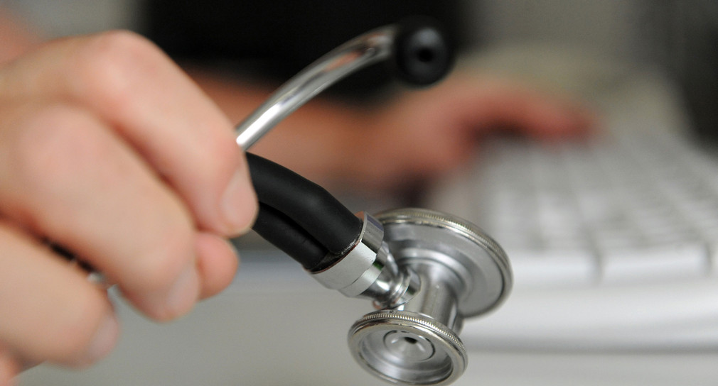 Ein Arzt hält in einem Behandlungszimmer in seiner Praxis ein Stethoskop in der Hand, mit der anderen Hand bedient er eine Computertastatur.. 
