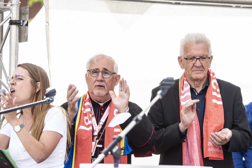 Ministerpräsident Winfried Kretschmann (rechts) und Bischof Dr. Gebhard Fürst (Mitte) klatschen, links eine Sängerin.