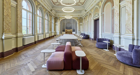 Lang gestreckter, historisch verzierter Raum mit hohen Rundbogenfenstern, Parkettboden und einer Orgel am Ende des Saals. Moderne Sitzmöbel und Tische bilden verschiedene Arbeits- und Aufenthaltsbereiche.