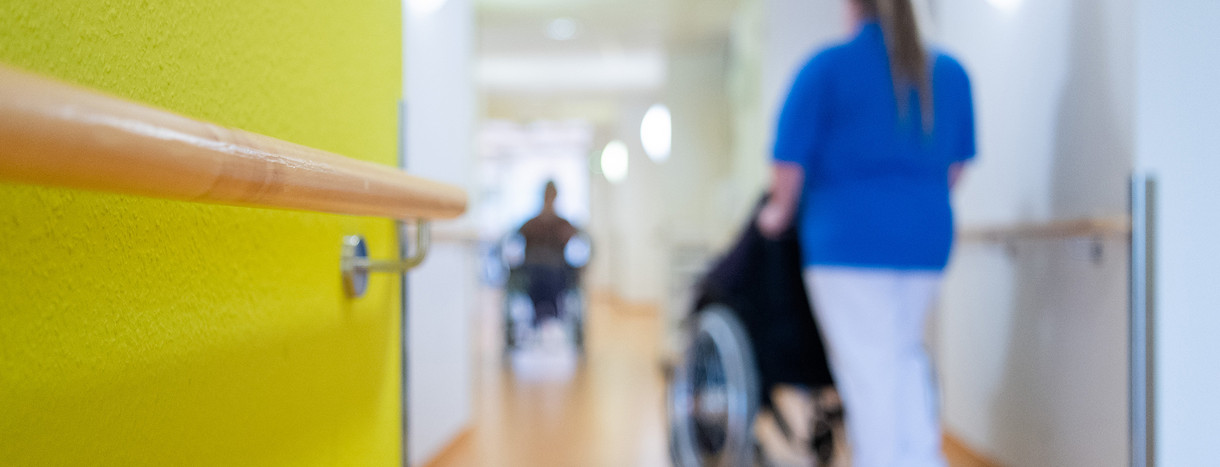 Eine Pflegerin schiebt eine Bewohnerin eines Pflegeheims in einem Rollstuhl über den Flur.