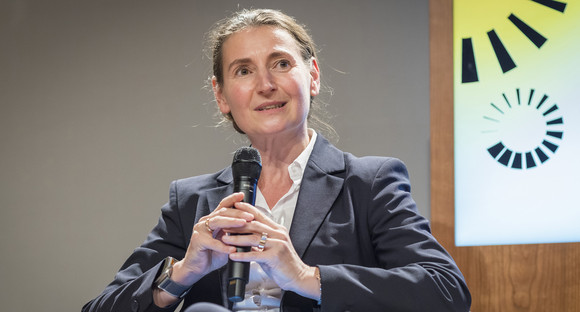 Susan-Stefanie Breitkopf sitzt auf dem Podium und blickt an der Kamera vorbei. Sie trägt eine weiße Bluse und ein dunkelgraues Jacket. In den Händen hält Frau Breitkopf ein Mikro. Im Hintergrund sieht man die Zeiträder des Key Visuals
