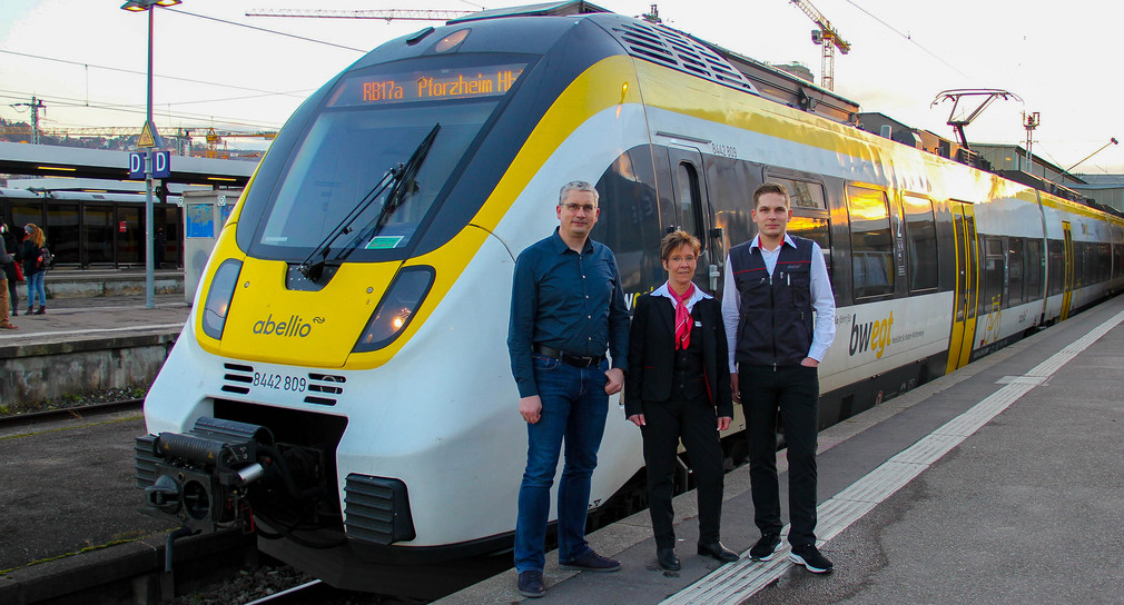 Drei Mitarbeitende von Abellio stehen vor einem gelb-weiß lackierten Zug und schauen in die Kamera.