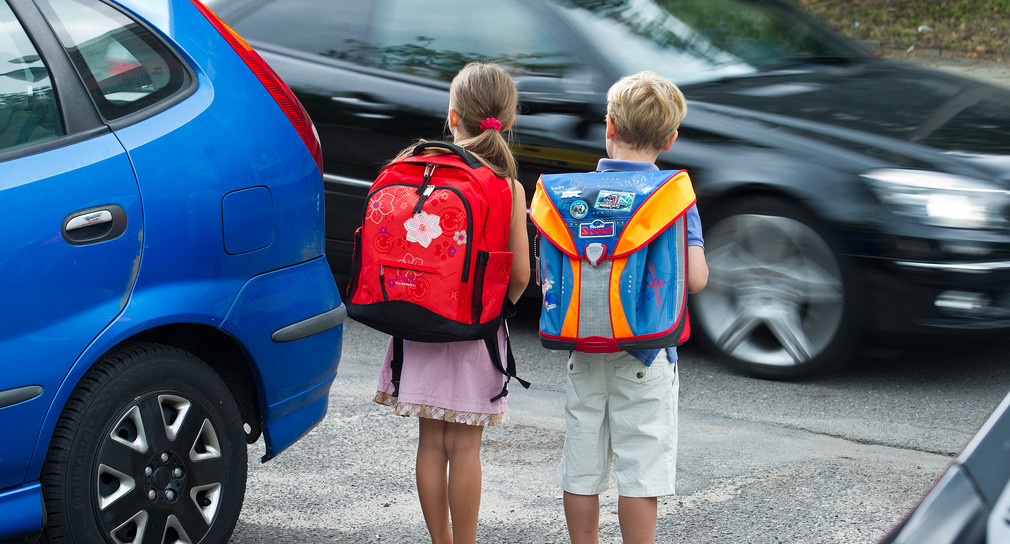 Zwei Schüler mit Schulranzen stehen an einer Straße um diese zu überqueren.