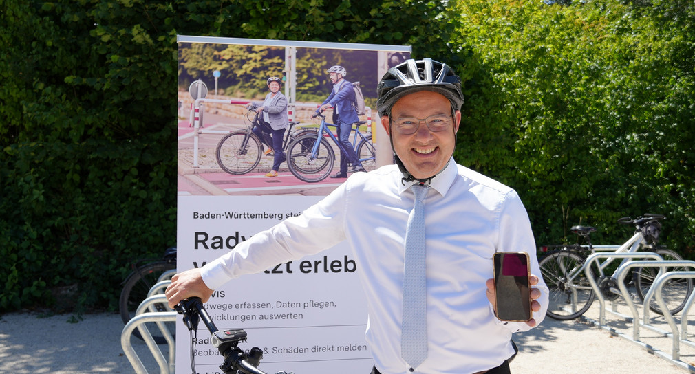 Staatssekretär Raimund Haser steht vor seinem Fahrrad mit einem Fahrradhelm auf dem Kopf und hält ein Handy in die Kamera.