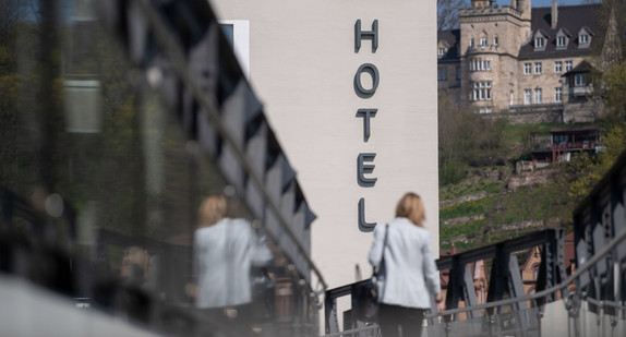 Eine Frau geht eine Fußgängerbrücke entlang, im Hintergrund ist ein Hotel zu sehen. (Bild: © picture alliance/Marijan Murat/dpa)