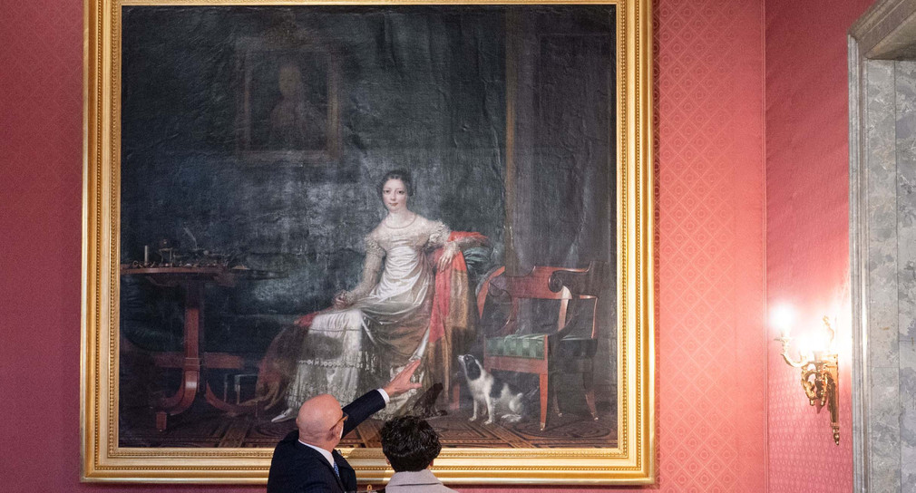 Ein Besucher und eine Besucherin einer Veranstaltung zur Enthüllung des Gemäldes „Königin Catharina von Württemberg in ihrem Arbeitszimmer im Neuen Schloss“ stehen im Empire-Zimmer des Neuen Schlosses in Stuttgart vor dem Gemälde.