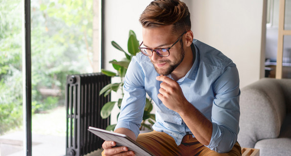 Ein Mann sitzt mit einem Tablet im Wohnzimmer (Bild: © StockRocket - stock.adobe.com)