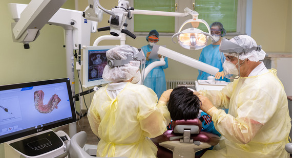 Ein Zahnarzt behandelt mit Visier, FFP3 Atemschutzmaske und Virenschutzkittel mit seinen Zahnmedizinischen Fachangestellten einen Patienten in seiner Zahnrztpraxis.