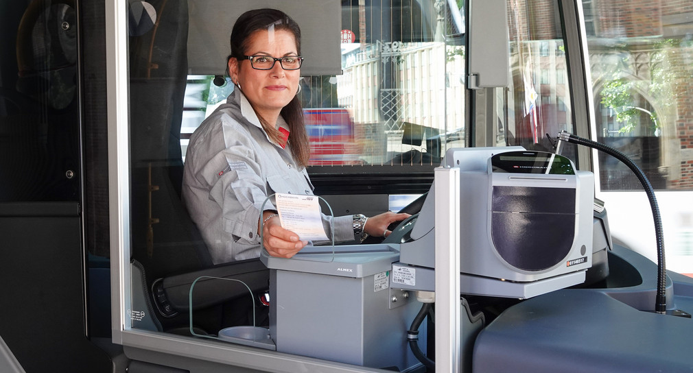 Eine Busfahrerin sitzt hinter dem Steuer und wird von den Fahrgästen mit einer Scheibe abgetrennt.