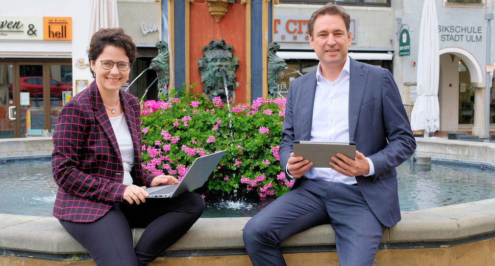 Die baden-württembergische Ministerin der Justiz Marion Gentges und der bayerische Staatsminister für Justiz Georg Eisenreich sitzen mit aufgeklappten Laptops an einem Brunnen. 