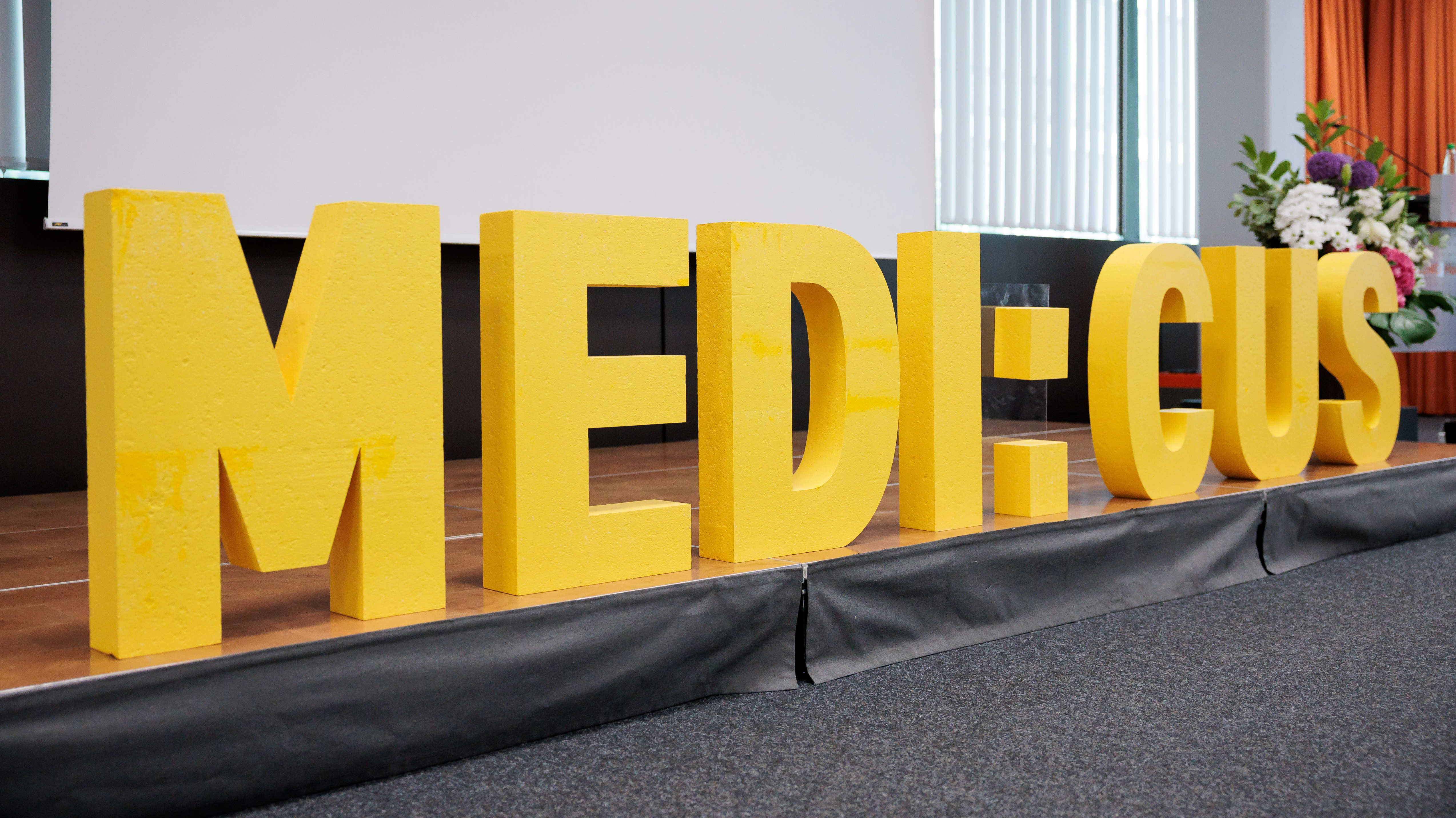Schriftzug &bdquo;MEDI:CUS&ldquo; auf einer B&uuml;hne