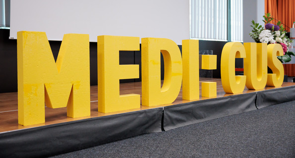 Schriftzug „MEDI:CUS“ auf einer Bühne