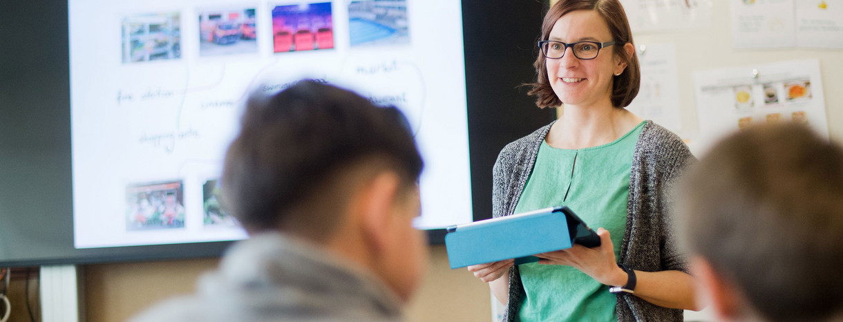 Eine Lehrerin mit einem Tablet in der Hand steht vor einer digitalen Tafel.