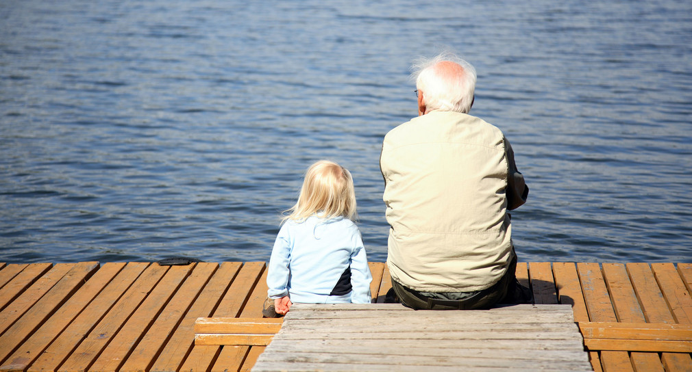 Ein kleines Kind und ein älterer Herr sitzen auf einem Steg an einem See.