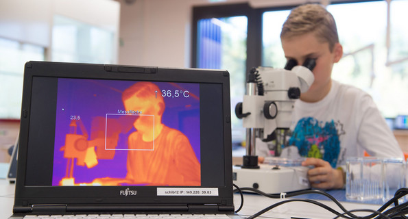 Ein Schüler sitzt an einem Mikroskop und wird dabei von einer Wärmekamera gefilmt.