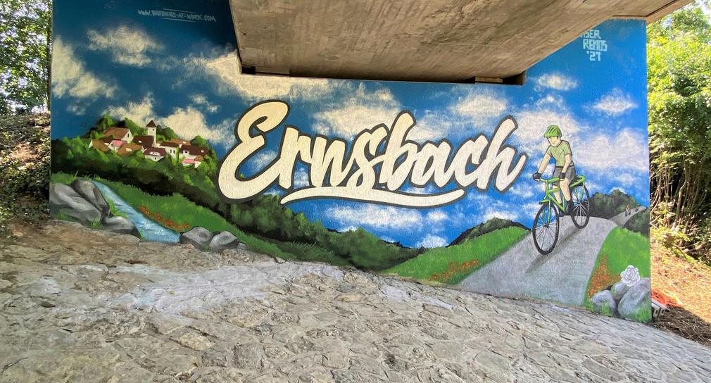 Ein Graffiti an einem Brückenpfeiler zeigt eine schöne grüne Landschaft mit einem Fahrradfahrer. Ein aufgesprühter Schriftzug weißt auf den Orts Ernsbach hin.
