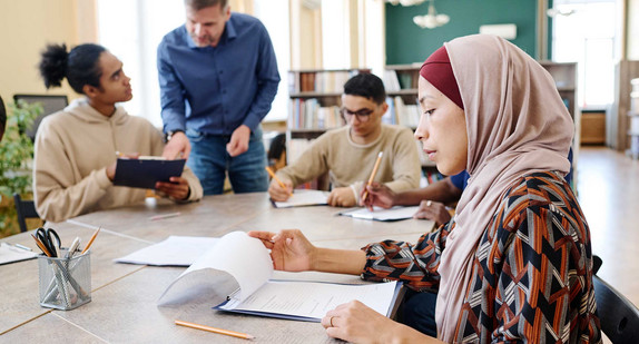 Eine muslimische Einwanderin sitzt mit anderen Personen am Tisch und schaut sich während des Englischunterrichts Blätter mit Grammatikaufgaben an.